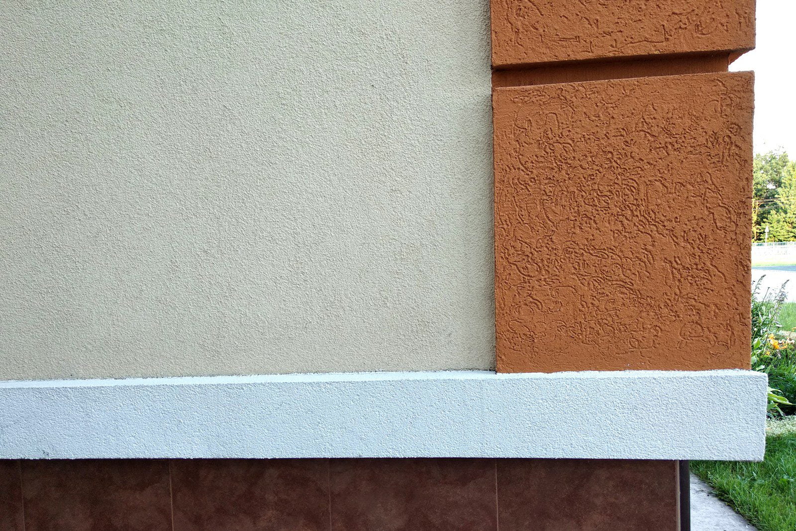 This image shows the corner of a building with an exterior wall finish. We can see a combination of different textures and colours.On the lower portion of the wall, there are brown tiles with a glossy, polished finish that creates a smooth appearance. Above that is a white ledge or molding, which seems to be made from a different, possibly lighter material with a rough texture that contrasts with the tiles below.The upper wall sections are coated with a sort of plaster or stucco. The left side of the upper wall displays a light beige colour with a slightly bumpy, uniform texture. On the right side, a column of the same finish but in a terracotta or rust colour protrudes slightly from the wall. The texture on this column is more pronounced and swirly, which indicates that a special technique was likely used during the stucco application to create this pattern.The wall's varied textures and colours may serve both aesthetic and functional purposes, providing not only visual interest but perhaps different levels of weather protection as well. The setting appears to be residential or possibly commercial, and it is daytime with plants visible in the background, indicating an outdoor environment.