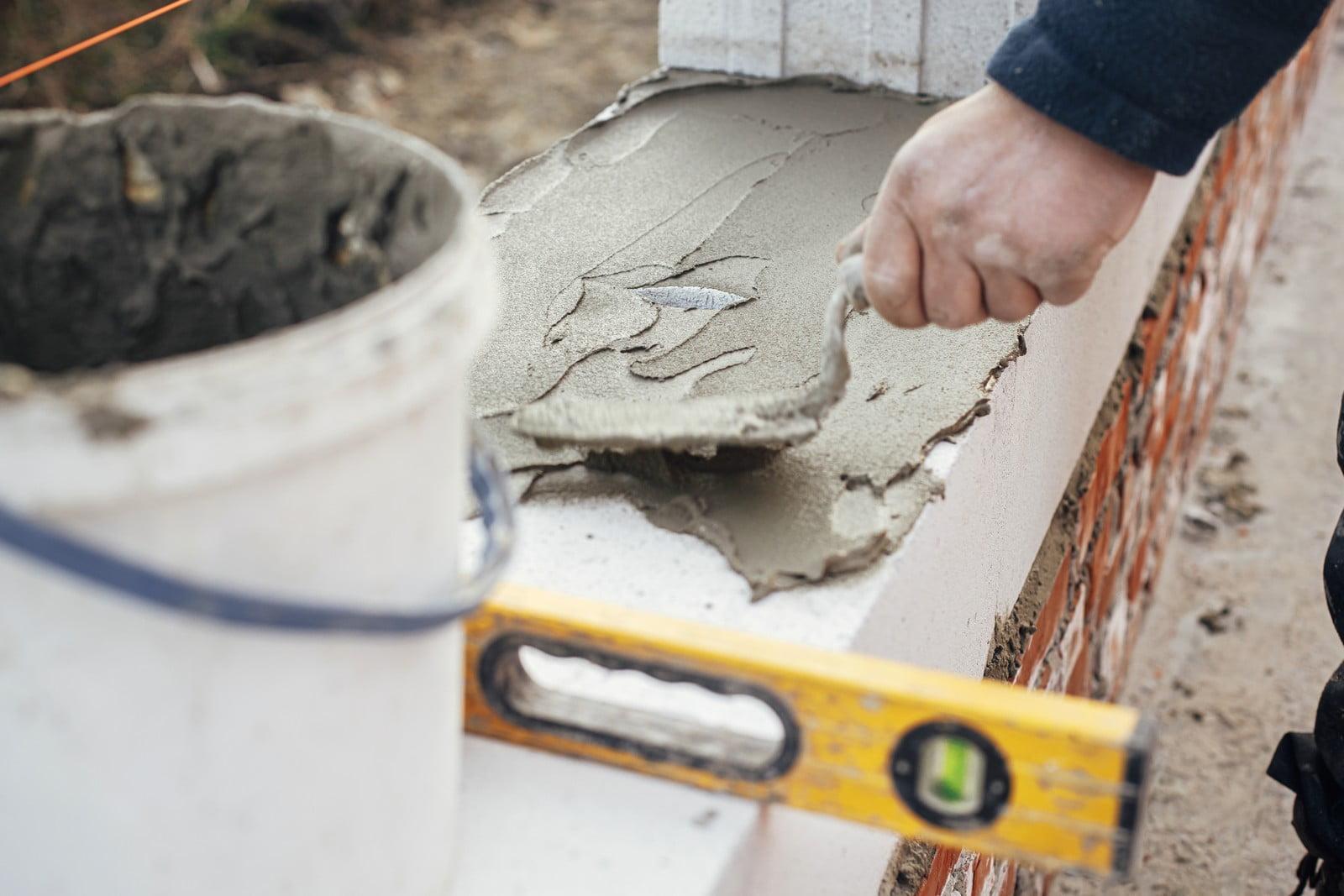 The image shows a person working with construction materials, likely in the process of building or repairing a structure. The person is spreading wet concrete or mortar onto the top of a brick or cinder block wall. In the foreground, there is a spirit level, which is used to ensure that the layers of the wall are level. To the left, there is a bucket filled with what appears to be the same wet concrete or mortar mixture being used on the wall. The focus of the photo is on the work being performed, with emphasis on the person's hand action applying the concrete and on the tools being used to ensure a level and even surface.