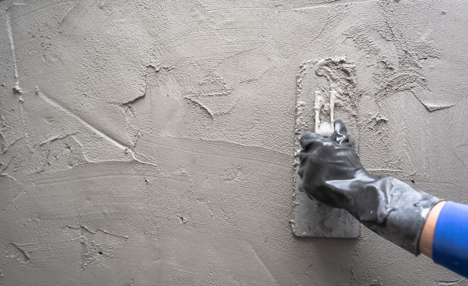 The image shows a hand in a black glove applying a layer of cement or plaster to a wall using a flat trowel. The wall already has some of the material applied, while the trowel is being used to smooth and spread the mixture evenly on the surface. It appears to be a construction or renovation project involving some sort of masonry or plastering work.