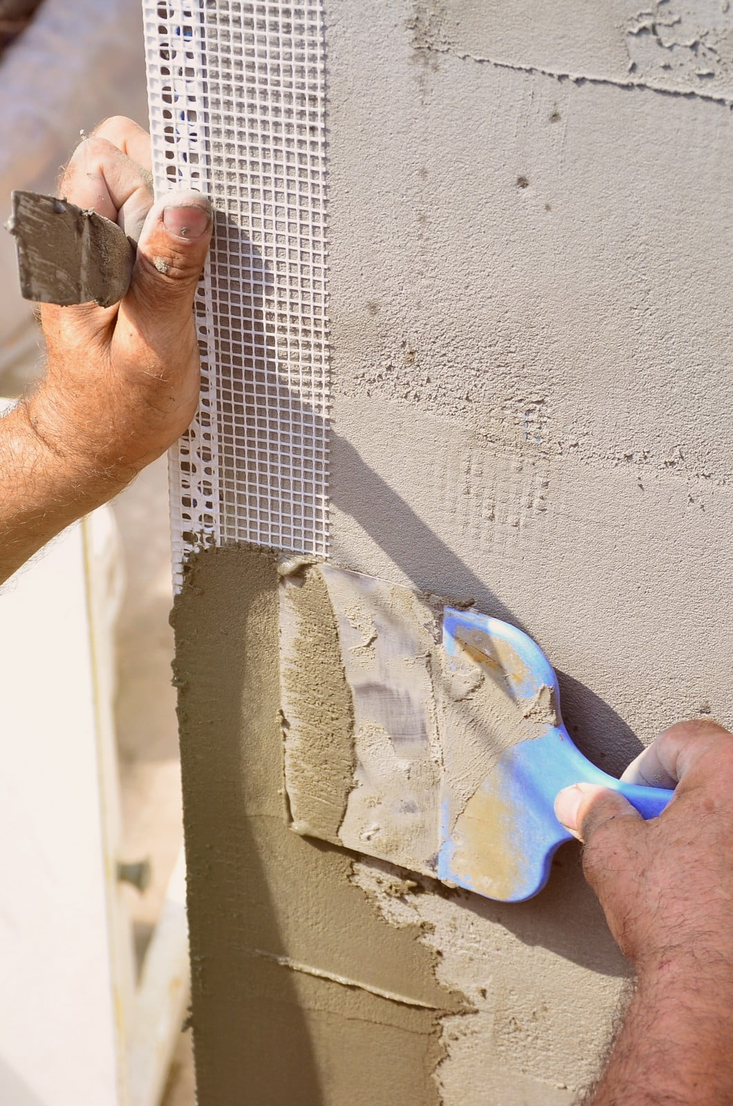 The image shows a person applying a plaster or stucco to a vertical surface. The person is using a trowel to smooth the material over a corner with mesh that will likely help the material adhere and prevent cracking. This is a common step in construction or renovation work to create a smooth, finished surface on walls or ceilings. The person is wearing a glove, and you can see their hands as they perform the work.