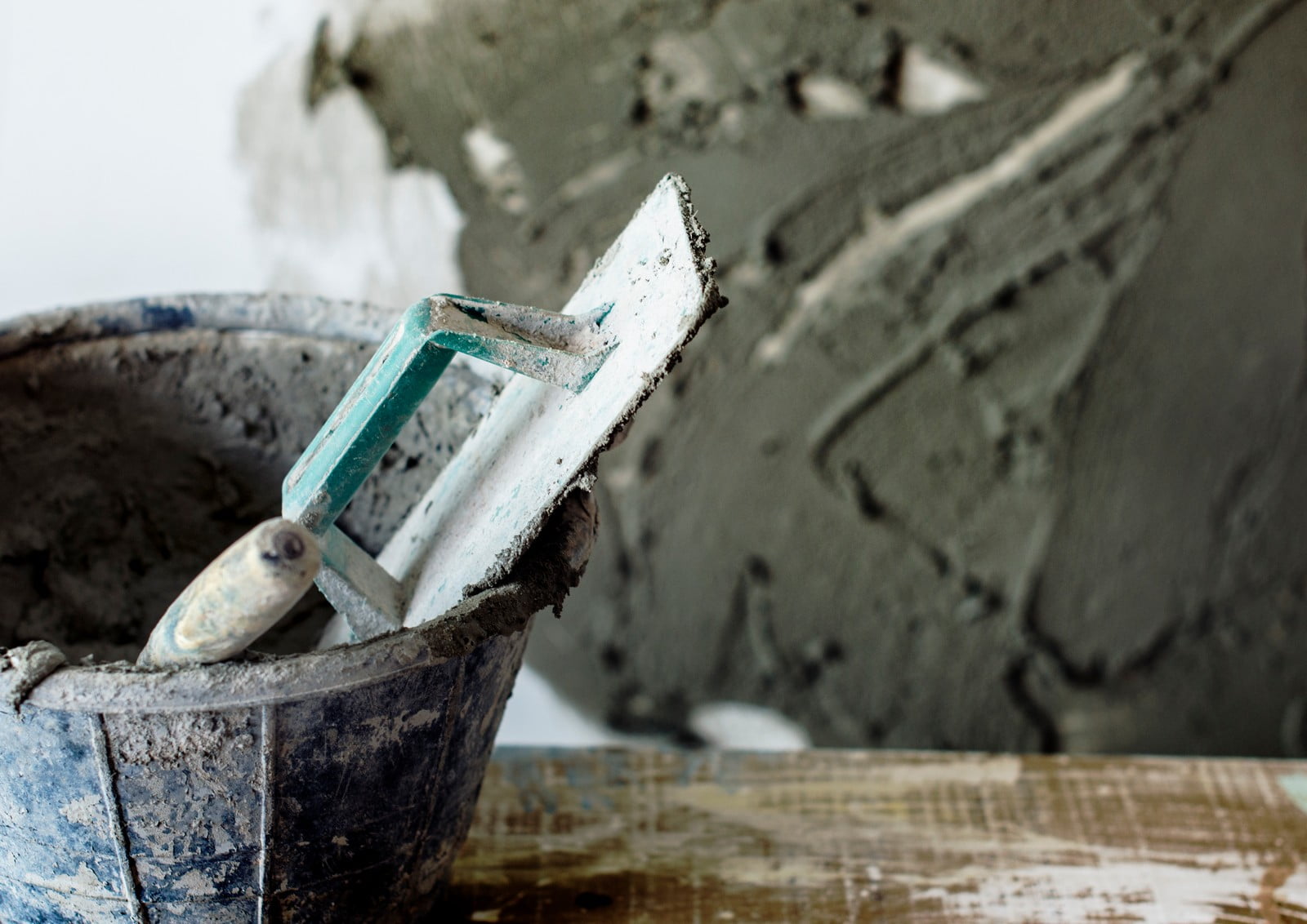 The image shows a close-up of construction tools and materials typically used for plastering or masonry work. In the foreground, there is a well-used plastering trowel with remnants of material on it, resting on the edge of a blue mortar mixing bucket, which also appears to contain some plaster or mortar mix. In the background, out of focus, is a wall with a fresh layer of plaster applied to it in a rough state before smoothing. The scene captures the dusty and messy environment often associated with construction and renovation work.